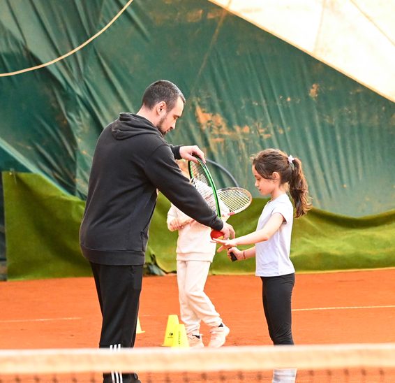 Antrenor care îi dă racheta unei fetițe de vârstă preșcolară, pe terenul de zgură
